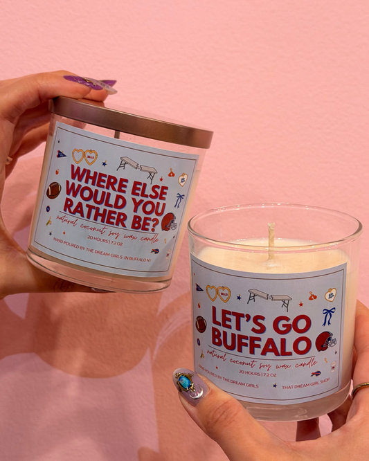 Two hands holding scented soy candles with humorous labels against a solid pink background. One candle reads 'WHERE ELSE WOULD YOU RATHER BE?' and the other reads 'LET'S GO BUFFALO'.