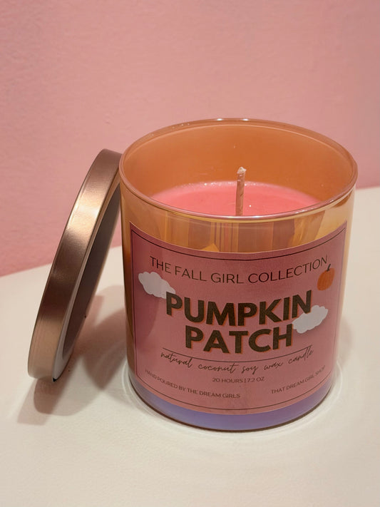 A lit jar candle with pink wax in a translucent peach-colored glass container, shown with its lid leaning beside it on a white surface against a pink wall. The front label reads 'Pumpkin Patch' and 'The Fall Girl Collection.