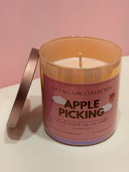 A 12 oz scented candle in a translucent amber glass jar with a pink label reading 'APPLE PICKING' and 'THE FALL GIRL COLLECTION.' The lid leans against the left side of the jar on a white surface against a pink background.