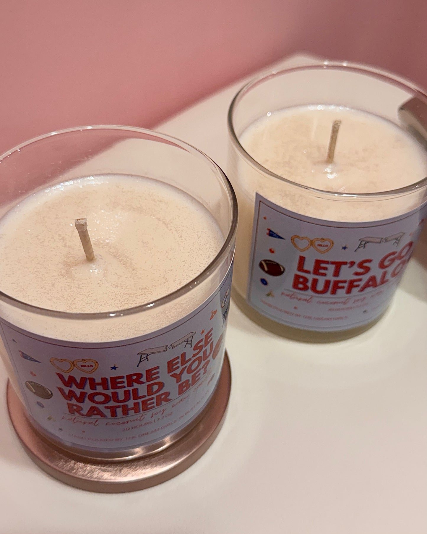 Two glass jar candles with white wax and wooden wicks, featuring humorous labels. The candle in the foreground reads 'WHERE ELSE WOULD YOU RATHER BET' with a rose gold coaster underneath, and the candle behind it reads 'LET'S GO BUFFALO' with a silver coaster underneath, set on a white surface against a pink background.