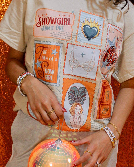 Person wearing a 'Show Girl' themed shirt with decorative elements, holding a disco ball against an orange sequin background.